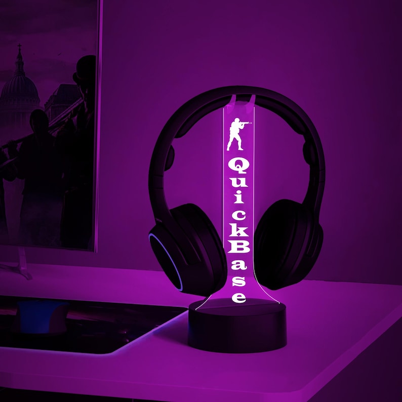 K&ouml;nnte beinhalten: Eine schwarz-wei&szlig;e 3D-gedruckte Lampe mit einer Gamer-Silhouette und dem Text "QuickBase" auf einem schwarzen Sockel. Die Lampe leuchtet in Lila und hat einen schwarzen Kopfh&ouml;rer darauf.