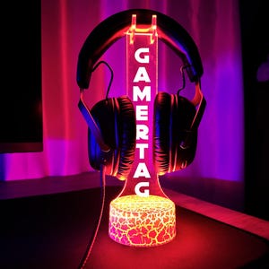 以下が含まれることがあります： 黒いゲーミングヘッドセットが、"GAMERTAG"の文字が白く光る透明なスタンドに置かれています。スタンドのベースはひび割れた溶岩のデザインで、照明は赤と紫です。