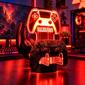 May include: A glowing red LED acrylic stand featuring a game controller outline with the text GAMER123. The stand holds a camouflage patterned wireless game controller and a black gaming headset, illuminated by a vibrant red light.
