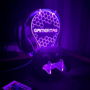 May include: A purple LED light stand with a honeycomb design and the word "GAMERTAG" in white. The stand holds a black video game controller and a pair of black headphones.