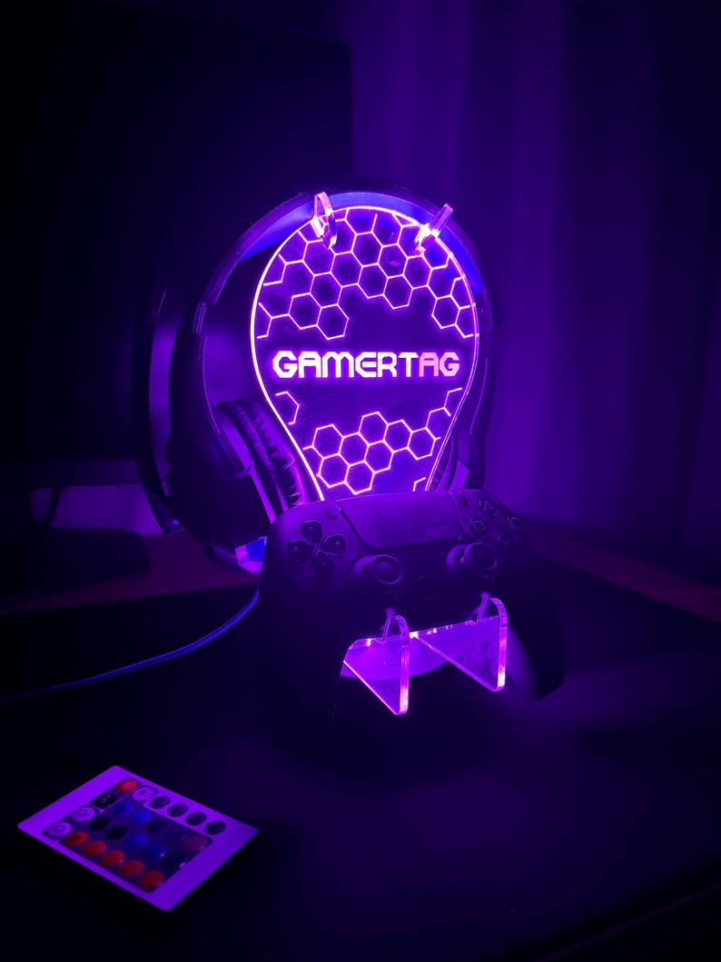 May include: A purple LED light stand with a honeycomb design and the word "GAMERTAG" in white. The stand holds a black video game controller and a pair of black headphones.
