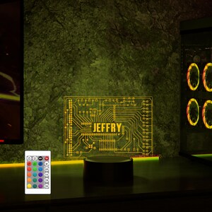 K&ouml;nnte beinhalten: Ein gelbes Schaltkreisdesign mit dem Namen "JEFFRY" in der Mitte. Das Design ist beleuchtet und steht auf einem schwarzen Sockel. Eine Fernbedienung mit mehreren farbigen Kn&ouml;pfen befindet sich im Vordergrund.