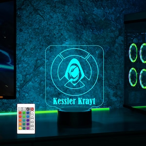 May include: A blue and white 3D printed night light with a hooded figure and a circle design. The light has a black base and a remote control with multiple color options. The light is personalized with the name "Kessler Krayt".