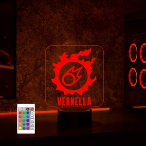 May include: A red LED night light featuring a stylised flame design with the name "VERNELLA" printed below. The light sits on a black base and comes with a remote control offering various colour options.