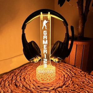 May include: A clear acrylic headset stand with a gamer silhouette and the text "GAMER123". The stand has a textured base and is lit from within, casting a warm glow.