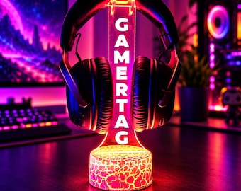 Może przedstawiać: Czarne słuchawki nauszne spoczywają na przezroczystym akrylowym stojaku z pionowym napisem GAMERTAG. Stojak posiada okrągłą podstawę z wzorem spękanej tekstury, podświetloną żywym czerwonym i pomarańczowym światłem.