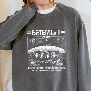 May include: Dark grey sweatshirt with the text "=ARTEMIS II= NASA" and a graphic of astronauts. The sweatshirt also has the text "Earth is rare. Treat it that way. AROUND THE MOON FOR ALL HUMANITY".