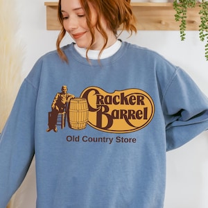 May include: A light blue sweatshirt featuring the "Cracker Barrel Old Country Store" logo in brown and yellow. The logo includes a man, a barrel, and the text "Cracker Barrel" and "Old Country Store".