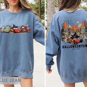 K&ouml;nnte beinhalten: Blaues Jeans-Sweatshirt mit Halloween-Motiv. Auf der Vorderseite sind Cartoon-Autos in Kost&uuml;men abgebildet, auf der R&uuml;ckseite eine Stadtszene mit "Halloweentown Est. 2006". Ein bequemes Langarm-Oberteil f&uuml;r die gruselige Jahreszeit.
