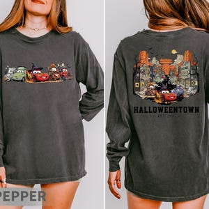 K&ouml;nnte beinhalten: Dunkelgraues Langarmshirt mit einem Halloween-Motiv von animierten Autos auf der Vorderseite. Die R&uuml;ckseite des Shirts zeigt eine detaillierte Illustration einer Stadt mit dem Text "Halloweentown" und "Est. 2003".
