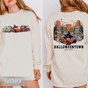 K&ouml;nnte beinhalten: Elfenbeinfarbenes Langarmshirt mit einem Halloween-Motiv von Zeichentrickautos auf der Vorderseite. Auf der R&uuml;ckseite des Shirts befindet sich eine detaillierte Illustration einer Stadt mit dem Text "Halloweentown Est. 2006".