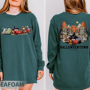 K&ouml;nnte beinhalten: Seegr&uuml;nes Langarm-Sweatshirt mit einem Halloween-Motiv von animierten Autos auf der Vorderseite. Auf der R&uuml;ckseite des Sweatshirts befindet sich eine detaillierte Illustration einer gruseligen Stadt mit dem Text "Halloweentown Est. 2006".