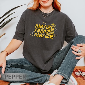 May include: Dark gray t-shirt with the word "AMAZE" in yellow, repeated three times with a planet and star graphic. The shirt is a relaxed fit and is paired with blue jeans.