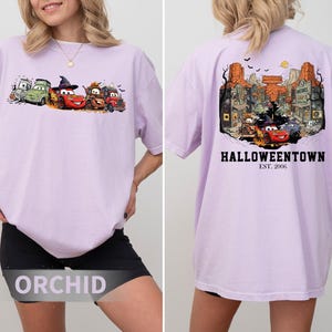 K&ouml;nnte beinhalten: Helllila T-Shirt mit einem Halloween-Motiv von Zeichentrickautos auf der Vorderseite. Die R&uuml;ckseite des Shirts zeigt eine detaillierte Illustration einer Stadt mit dem Text "Halloweentown" und "Est. 2006".