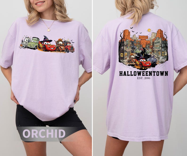 K&ouml;nnte beinhalten: Helllila T-Shirt mit einem Halloween-Motiv von Zeichentrickautos auf der Vorderseite. Die R&uuml;ckseite des Shirts zeigt eine detaillierte Illustration einer Stadt mit dem Text "Halloweentown" und "Est. 2006".