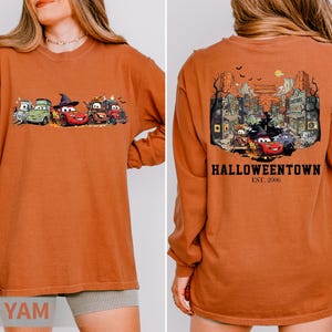 K&ouml;nnte beinhalten: Gebrannt orangefarbenes Langarmshirt mit einem Halloween-Motiv von animierten Autos. Auf der Vorderseite sind Autos in Kost&uuml;men zu sehen, w&auml;hrend die R&uuml;ckseite eine gruselige Stadtszene mit dem Text "Halloweentown Est. 2006" zeigt.
