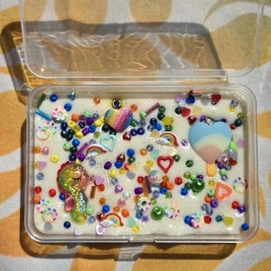 May include: A clear plastic container filled with white slime. The slime is decorated with colorful beads, rainbow sprinkles, and small charms, including a rainbow heart, a seahorse, and a popsicle.