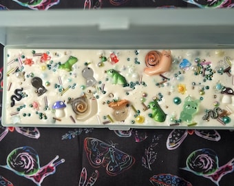 May include: A white rectangular box with a clear lid, filled with a white substance and decorated with various small, colorful figurines, including snails, frogs, and mushrooms.