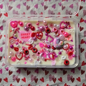 May include: A white wax melt with a variety of pink and red hearts, beads, and sprinkles. The wax melt is in a clear plastic container and says "Sweet Heart" on a pink heart.