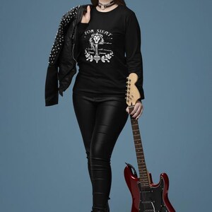 Op de afbeelding: Een vrouw draagt een zwart shirt met lange mouwen met een witte leeuw afbeelding en de tekst "FOR SIERT" en houdt een rode elektrische gitaar vast. Ze draagt ook een zwart leren jack met zilveren studs.