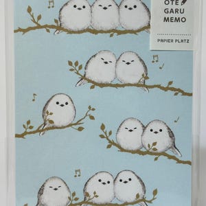 Peut inclure: Un bloc-notes avec un fond bleu clair orné d'illustrations d'oiseaux blancs perchés sur des branches brunes. Les oiseaux ont des yeux noirs et sont disposés par paires et en groupes. Le texte "OTE GARU MEMO" et "PAPIER PLATZ" est visible.