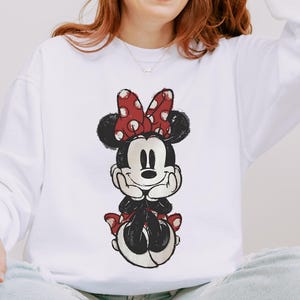 May include: White sweatshirt featuring a distressed graphic of Minnie Mouse. The cartoon character is depicted in black, red, and white, with a red polka-dot bow. The sweatshirt is a long-sleeved, crew neck style.