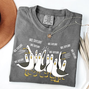 May include: A gray Comfort Colors t-shirt featuring a cartoon of seagulls with the text "MS. TAYLOR!". The seagulls are white with black wings and yellow feet. A gold necklace is draped on the shirt.