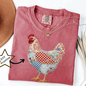 May include: A red Comfort Colors t-shirt with a patchwork chicken design. The chicken has a red comb, floral, gingham, and blue patterned details. A silver necklace with a pendant is draped on the shirt. The shirt is labelled "S-P-CH".