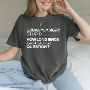 May include: Dark gray t-shirt with the text "GRUMPY. ANGRY. STUPID. HOW LONG SINCE LAST SLEEP, QUESTION?" in white. The shirt is a casual style, suitable for everyday wear.