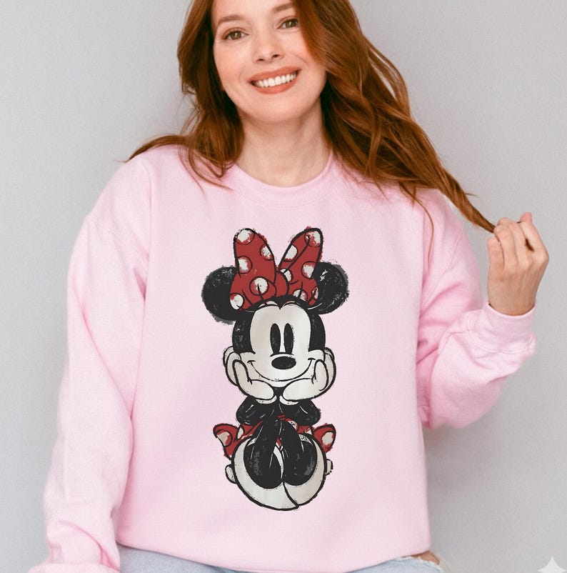 May include: A light pink sweatshirt featuring a Minnie Mouse graphic. The graphic shows Minnie Mouse in a seated pose, with a red bow with white polka dots, and a black and white outfit. The sweatshirt is a crew neck style.