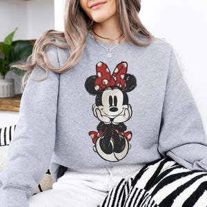 May include: A light gray sweatshirt featuring a Minnie Mouse graphic. The graphic shows Minnie Mouse in a seated pose, with a red bow with white polka dots, black ears, and a black outfit. The sweatshirt is a casual style.