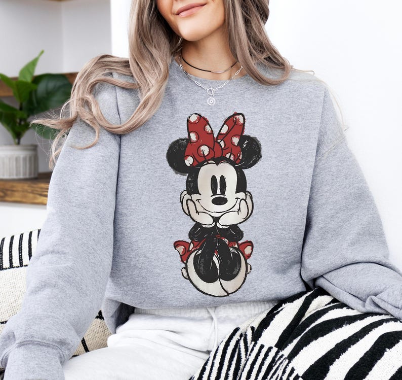 May include: A light gray sweatshirt featuring a Minnie Mouse graphic. The graphic shows Minnie Mouse in a seated pose, with a red bow with white polka dots, black ears, and a black outfit. The sweatshirt is a casual style.
