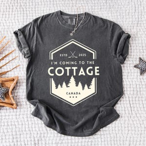 May include: Dark gray t-shirt with a white graphic that reads "I'M COMING TO THE COTTAGE" with a hockey stick design and the word "CANADA". The shirt has a crew neck and short sleeves. A silver necklace is visible.