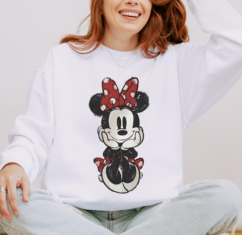 May include: White sweatshirt featuring a distressed graphic of Minnie Mouse. The cartoon character is depicted in black, red, and white, with a red polka-dot bow. The sweatshirt is a long-sleeved, crew neck style.