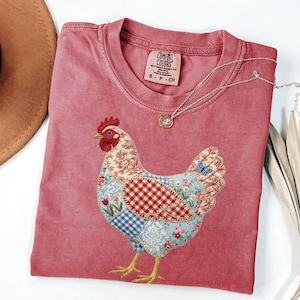 Peut inclure: T-shirt Comfort Colors rouge avec un motif de poulet patchwork. Le poulet est composé de motifs floraux, à carreaux et cachemire. Un collier argenté avec un pendentif repose sur le t-shirt. Un chapeau marron est visible.