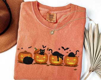 May include: Orange Comfort Colors t-shirt featuring a Halloween design. The graphic shows four jack-o'-lanterns with black cats inside. Bats and stars are also included in the design. A gold necklace and a brown hat are also visible.