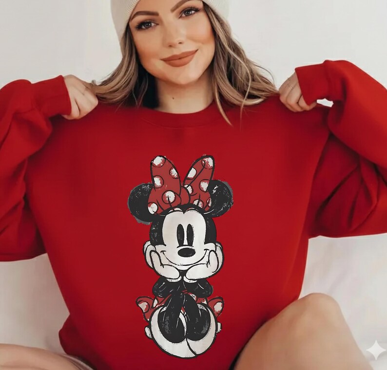 May include: A red sweatshirt featuring a Minnie Mouse graphic. The graphic depicts Minnie Mouse with a red polka dot bow, black ears, and a smiling face. The sweatshirt is a solid red color.