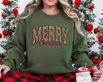 May include: An olive green sweatshirt with the words "MERRY Christmas" in a leopard print design. The word "Christmas" is in a red script font. The person is wearing red and black plaid pajama pants.