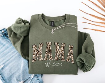May include: An olive green sweatshirt with the word "MAMA" in a leopard print design. The text "est. 2025" is below the word "MAMA". A silver necklace with a pendant is on the sweatshirt. A pair of blue jeans is on the left.