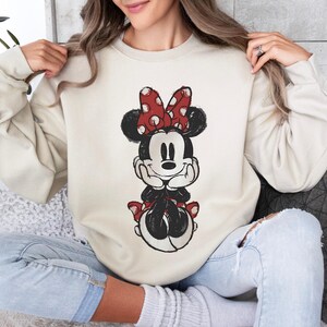 May include: Beige sweatshirt featuring a distressed Minnie Mouse graphic. Minnie is depicted with a red polka dot bow, black ears, and a black and white outfit. The sweatshirt has long sleeves and a crew neck.