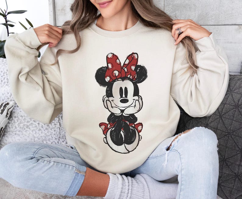 May include: Beige sweatshirt featuring a distressed Minnie Mouse graphic. Minnie is depicted with a red polka dot bow, black ears, and a black and white outfit. The sweatshirt has long sleeves and a crew neck.