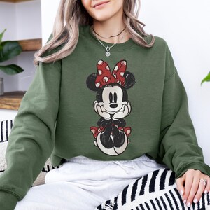 May include: Olive green sweatshirt featuring a distressed Minnie Mouse graphic. The graphic shows Minnie Mouse with a red polka dot bow and matching accents. The sweatshirt is a relaxed fit and has long sleeves.