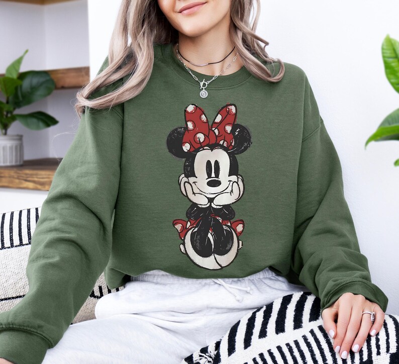 May include: Olive green sweatshirt featuring a distressed Minnie Mouse graphic. The graphic shows Minnie Mouse with a red polka dot bow and matching accents. The sweatshirt is a relaxed fit and has long sleeves.