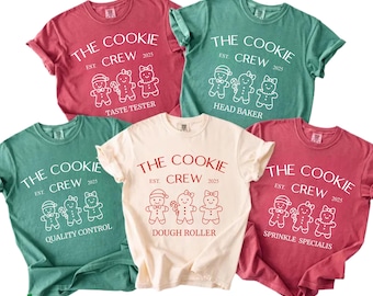 May include: Five short-sleeved t-shirts in shades of red, green, and cream. Each shirt features the text "THE COOKIE CREW EST. 2025" with gingerbread people and job titles like "Taste Tester" and "Dough Roller".
