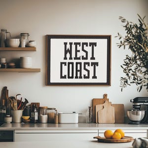 May include: A framed black and white pixel art print with the text "WEST COAST" hanging on a white wall in a kitchen. The kitchen has wooden shelves with dishes and jars, a wooden cutting board, and a stand mixer.