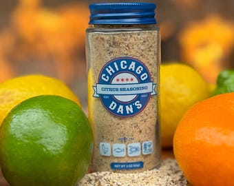 May include: A glass jar with a blue lid filled with a citrus seasoning blend. The label reads "Chicago Dan's Citrus Seasoning Est. 2021" with three stars and the text "Best On" with icons of a chicken, fish, and crab. The jar is sitting on a wooden surface with lemons, limes, and oranges around it.