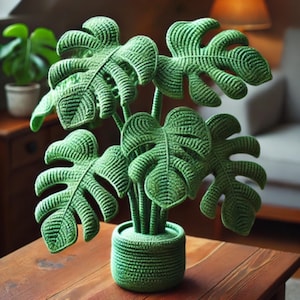 Puede incluir: Una planta de monstera de crochet verde con varias hojas en una maceta de crochet verde. La planta est&aacute; sobre una mesa de madera.