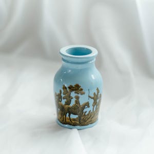 May include: A pale blue ceramic vase with a detailed brown and gold relief design. The relief shows figures on horseback and in a landscape. The vase has a cylindrical shape with a flared rim.