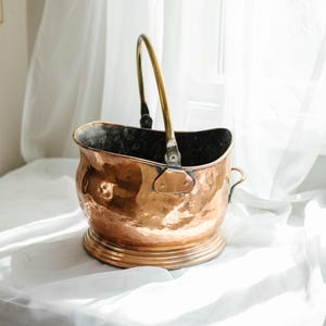 May include: A copper bucket with a brass handle. The bucket has a rounded body with a decorative base and a curved handle. The metal has a warm, reddish-brown hue, and the handle is a contrasting gold colour. The bucket is likely vintage.