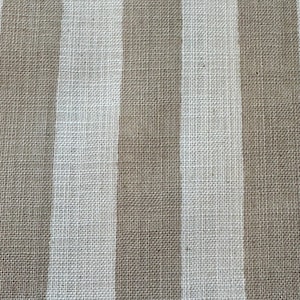 May include: A woven fabric featuring a repeating pattern of vertical stripes in alternating shades of light beige and off-white. The material displays a distinct, coarse, and textured weave characteristic of natural fibers, creating a uniform surface.